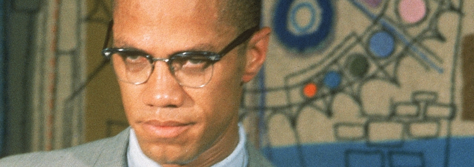 Color image of Malcolm X in suit and tie in front of multicolored abstract image.